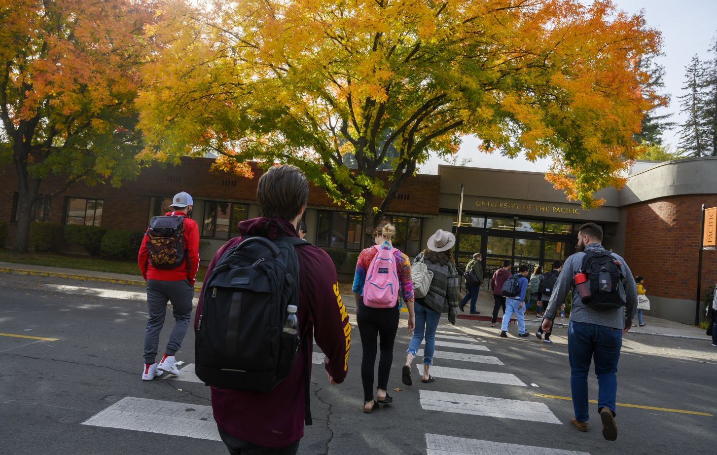 Students wearing backpacks cross the street during a fall day.