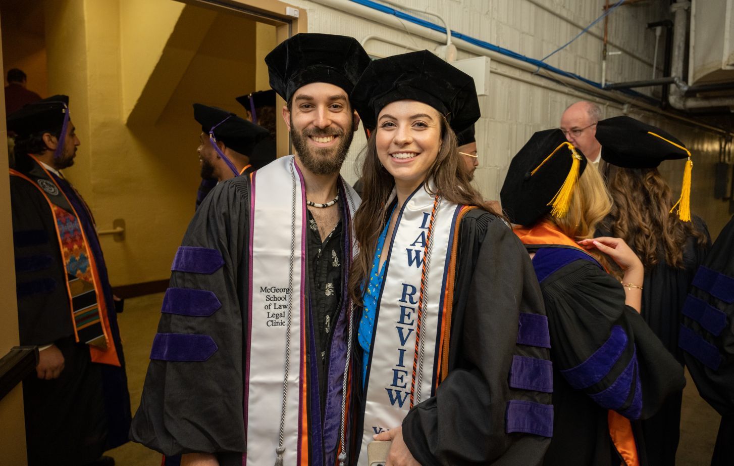 One male individual and one female individual wearing graduation regalia pose for a photo in the basement of an event venue
