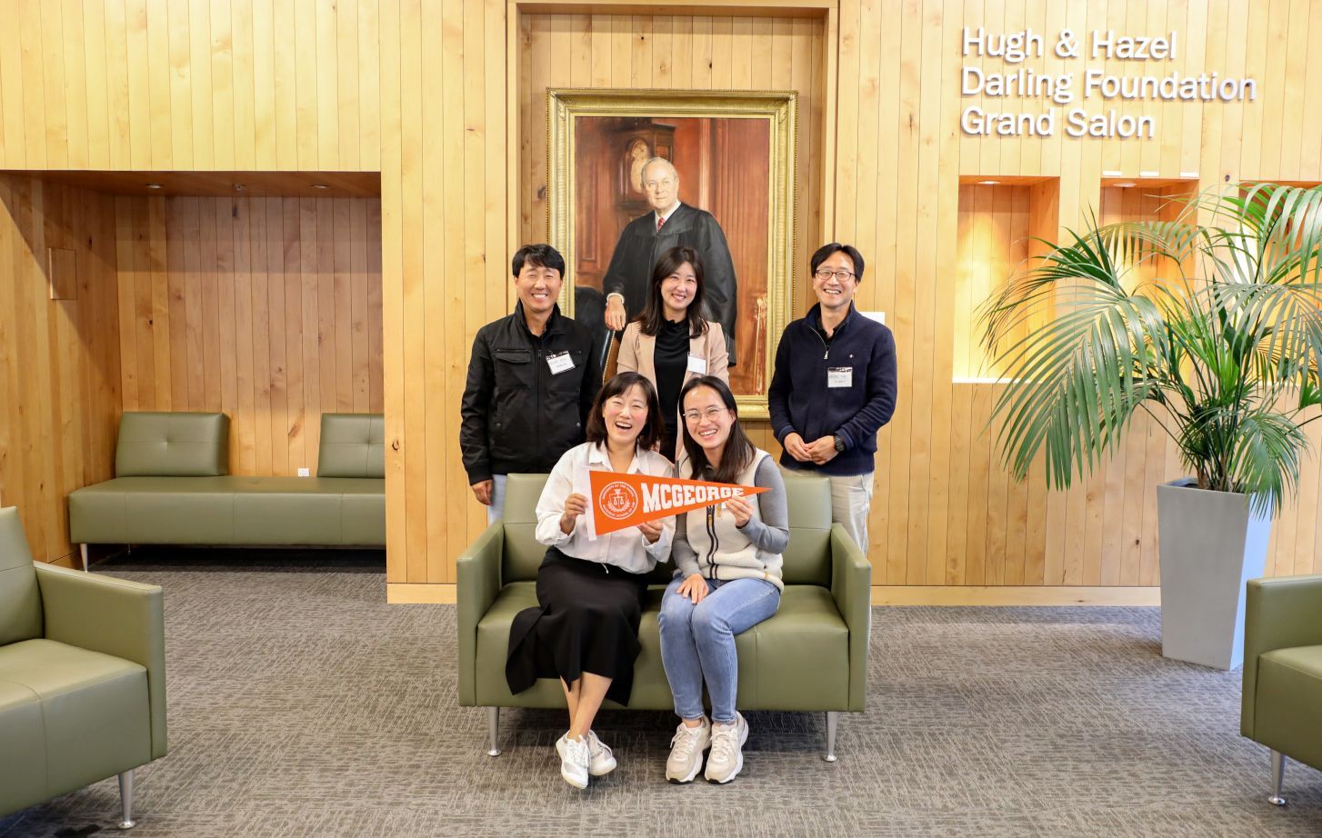 Five Korean individuals pose for a photo. Three are standing, and two are sitting and holding an orange pennant.