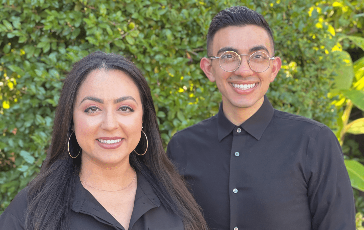 Two individuals wearing black pose for a photo in front of shrubbery