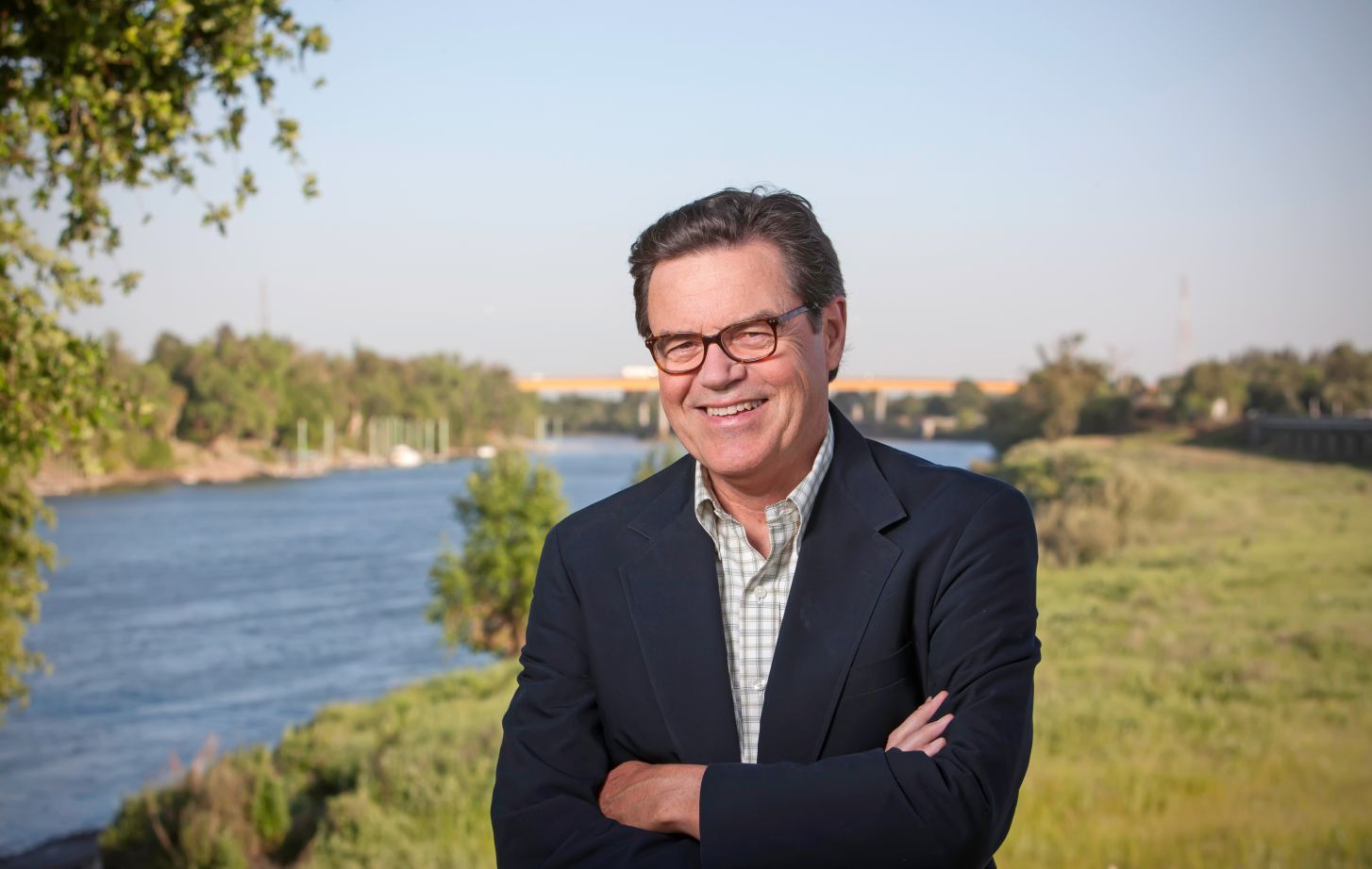 A man in a suit stands smiling in front of a river.