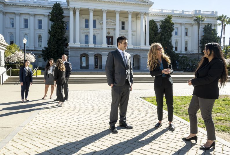 A group of people in suits converse with one another in front of the the California State Capitol Building